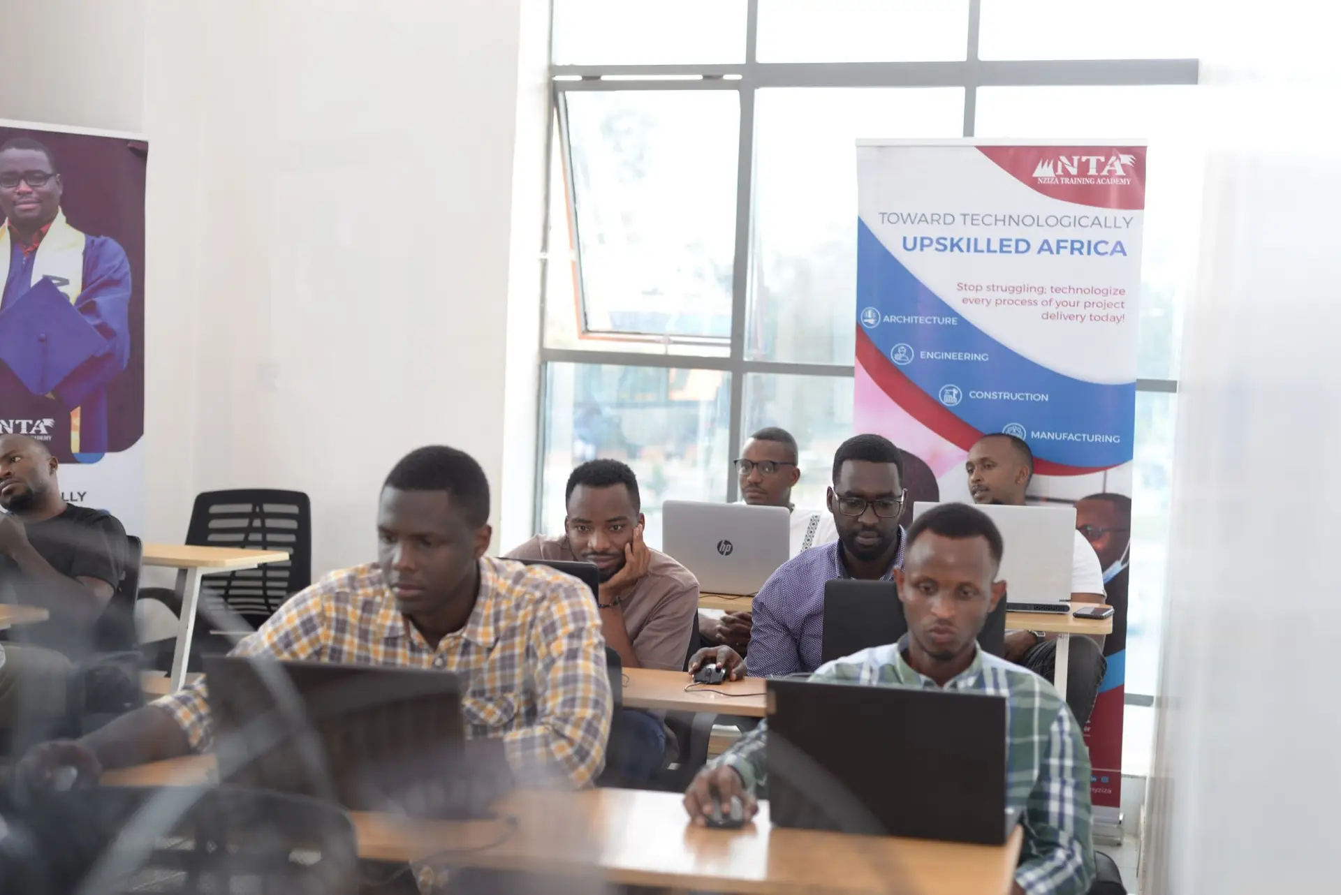 Focused group of African male professionals working on laptops in a bright classroom with educational banners, including one promoting technological upskilling